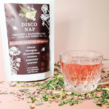 A clear glass vintage tea cup filled with a hibiscus pink-colored herbal tea, alongside a package labeled 'DISCO NAP' with botanical illustrations and text describing the tea's ingredients and purpose