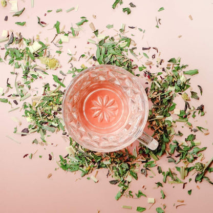 Aerial view of a vintage glass tea cup filled with a hibiscus pink-hued herbal tea surrounded by scattered loose-leaf tea against a light pink background