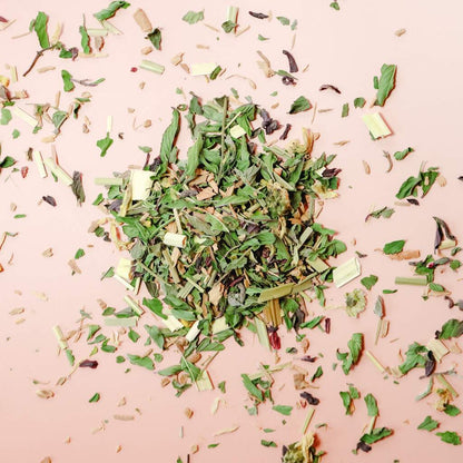 A pile of loose-leaf herbal tea of hibiscus, cinnamon, peppermint, lemongrass, and chyrsanthemum flowers against a light pink background