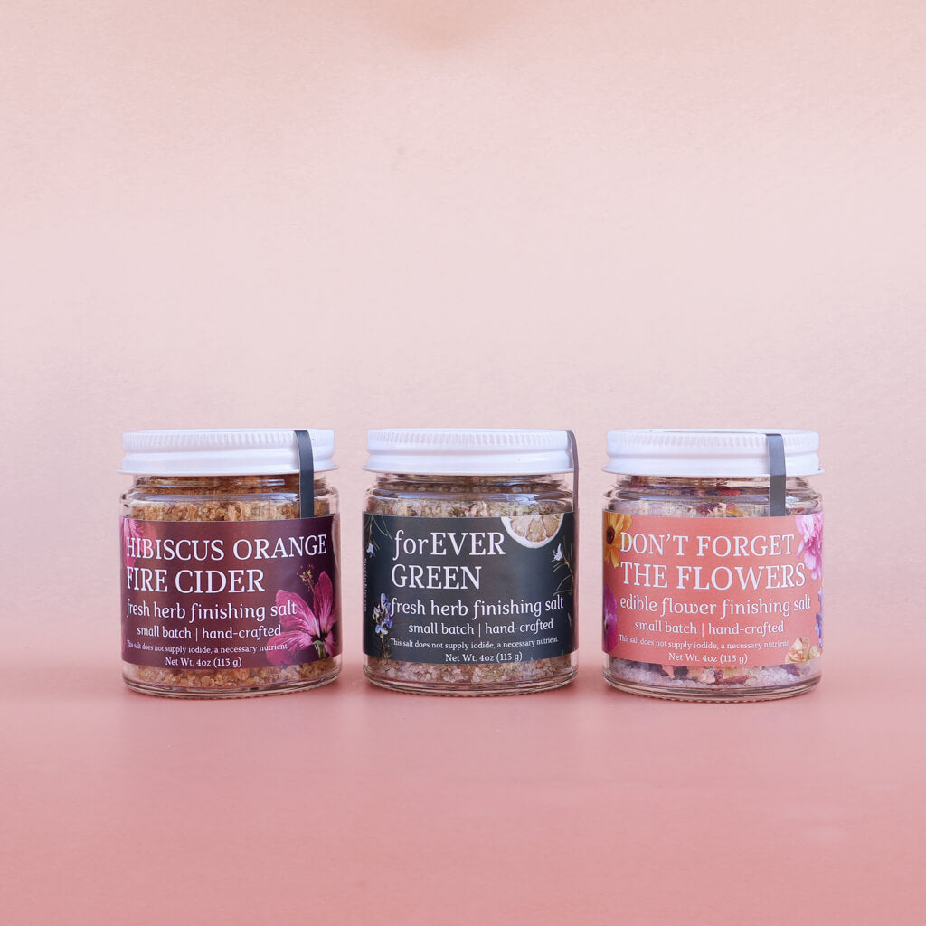 Three jars of 'Fire Cider', 'For EVER Green', and 'Don't Forget The Flowers' fresh herb finishing salts on a pink background