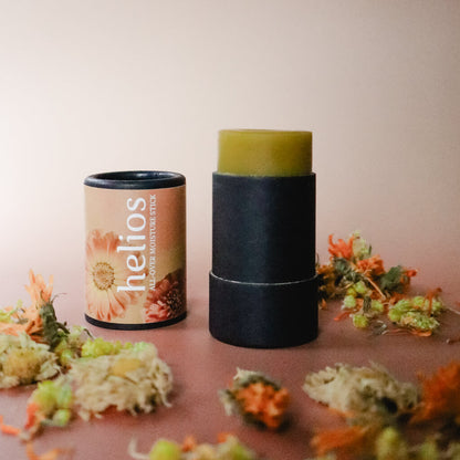 An opened black paper tube of Helios All-Over Moisture Stick herbal salve with a peach-colored floral label surrounded by dried calendula, helichrysum, and chrysanthemum, against a light pink background
