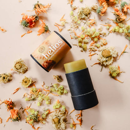 A solid golden-colored stick of Helios All-Over Moisture Salve, with its packaging beside it, surrounded by dried calendula and helichrysum flowers.
