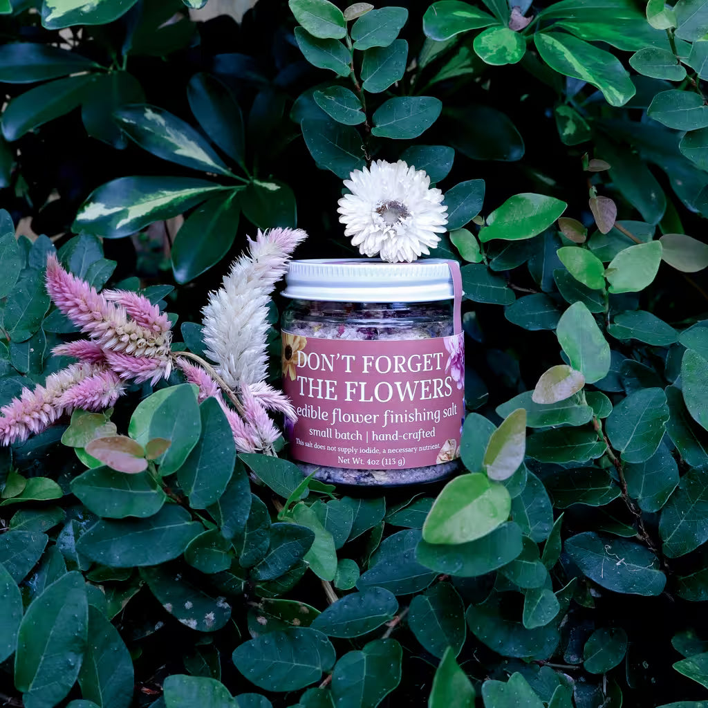 glass jar of herb infused salt with a label that reads 'Don't Forget the Flowers Edible Flower Finishing Salt' in a natural setting with lush greenery and dried flowers as decoration