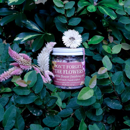 glass jar of herb infused salt with a label that reads 'Don't Forget the Flowers Edible Flower Finishing Salt' in a natural setting with lush greenery and dried flowers as decoration