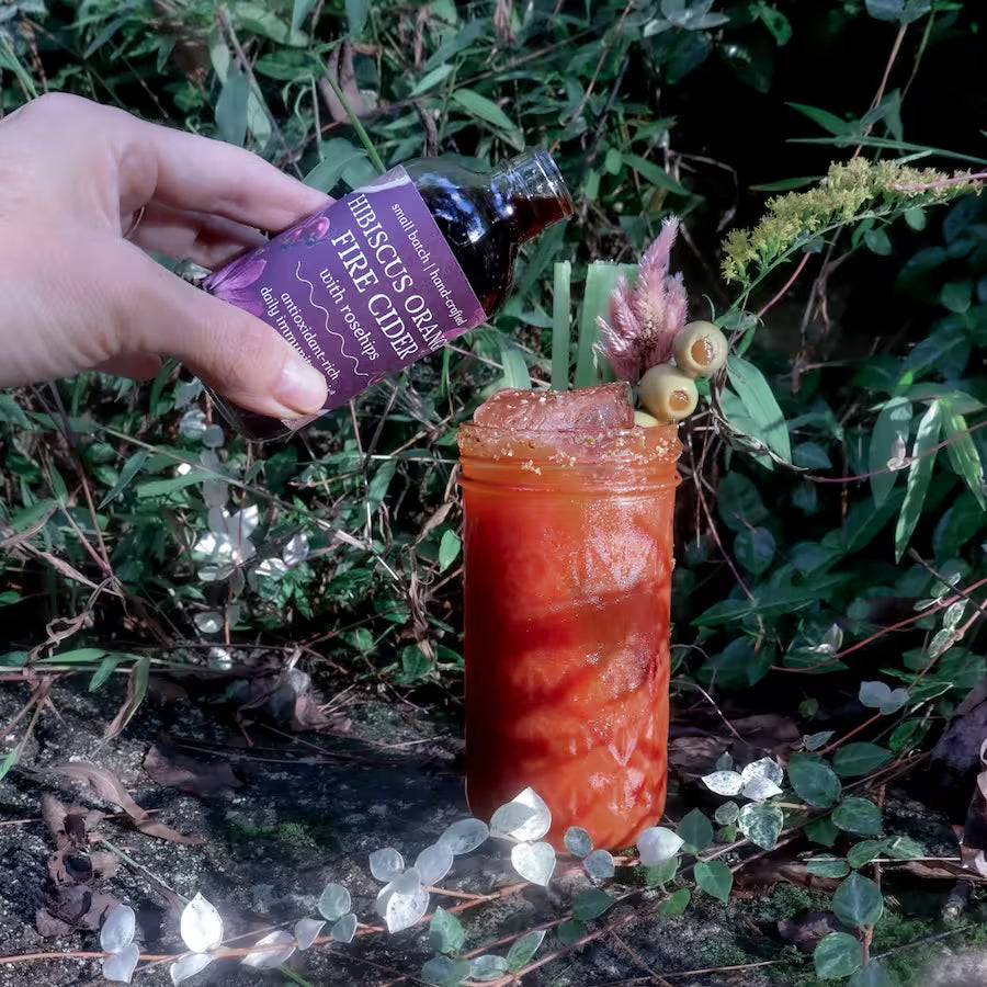 a Bloody Mary cocktail in a tall slender glass garnished with celery, a dried pink flower, green olives speared with a stem of goldenrod flowers, and an herbal salt rim. A hand pours from a bottle of fire cider into the cocktail that sits on a moss-covered stone with greenery in the background.
