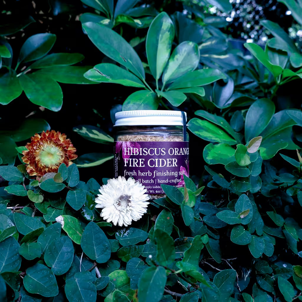 glass jar of herb infused salt with a label that reads 'Hibiscus Orange Fire Cider Fresh Herb Finishing Salt' in a natural setting with dried flowers as decoration