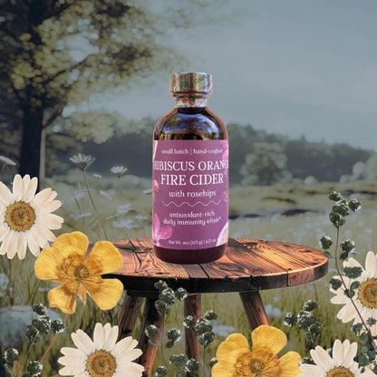 Bottle of HIBISCUS ORANGE FIRE CIDER with rosehips on a wooden stool with a nature-themed background.