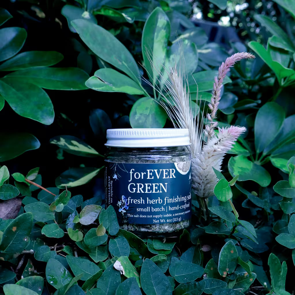 glass jar of herb infused salt with a label that reads 'Forever Green Fresh Herb Finishing Salt' in a natural setting with lush greenery and dried flowers as decoration