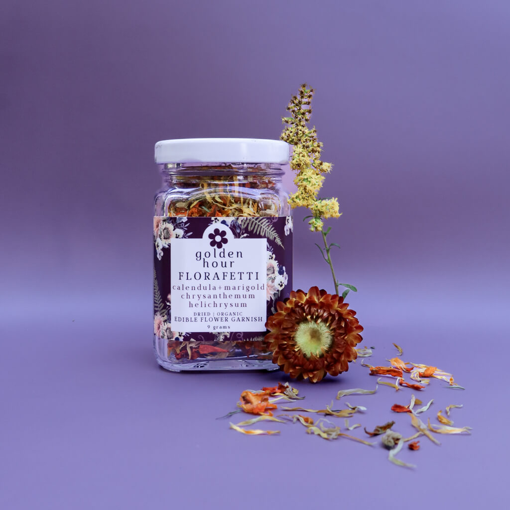 A glass jar of Golden Hour Florafetti Dried Edible Flower Garnish with Calendula, Marigold, and Chrysanthemum against a purple background with dried flower decorations.