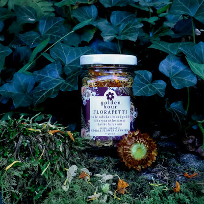 A glass jar of Golden Hour Florafetti Dried Edible Flower Garnish with Calendula, Marigold, and Chrysanthemum on a mossy rock with moody greenery in the background and flower confetti sprinkled about.
