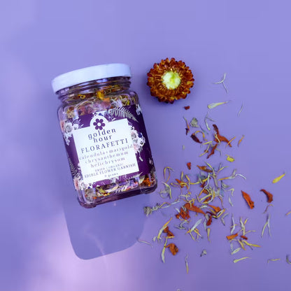 A glass jar of Golden Hour Florafetti Dried Edible Flower Garnish with Calendula, Marigold, and Chrysanthemum against a purple background with dried flower decorations.