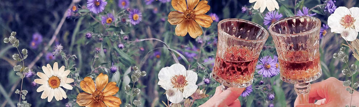 Two hands holding two glasses filled with pink herbal elixirs against a botanical background with purple, yellow, and white flowers