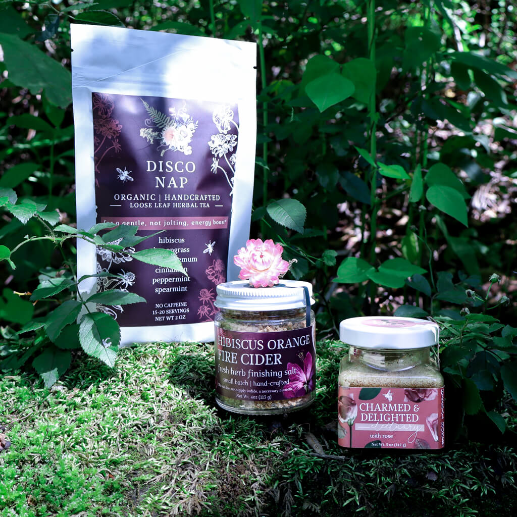 our bestsellers (a bag of herbal tea, a finishing salt, and an herbal honey) sitting on a mossy rock with greenery in the background
