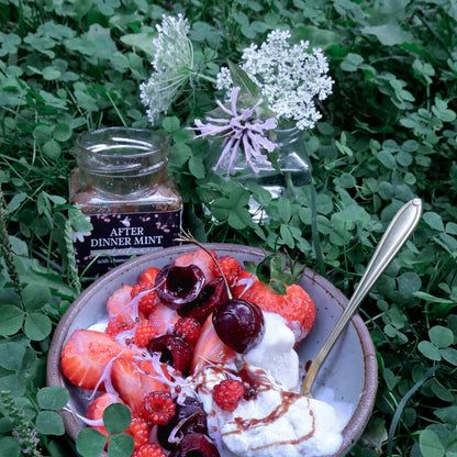 bowl of vanilla ice cream with red berries drizzled with mint chocolate honey next to a jar of After Dinner Mint Herbal Honey with greenery and flowers in the background