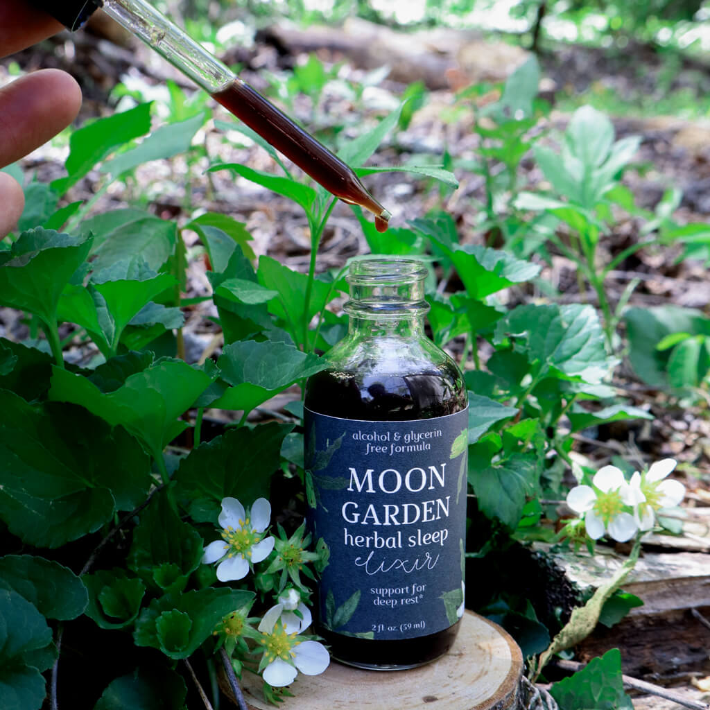 Bottle of 'Moon Garden Herbal Sleep Elixir' with a dropper over green plants and flowers.