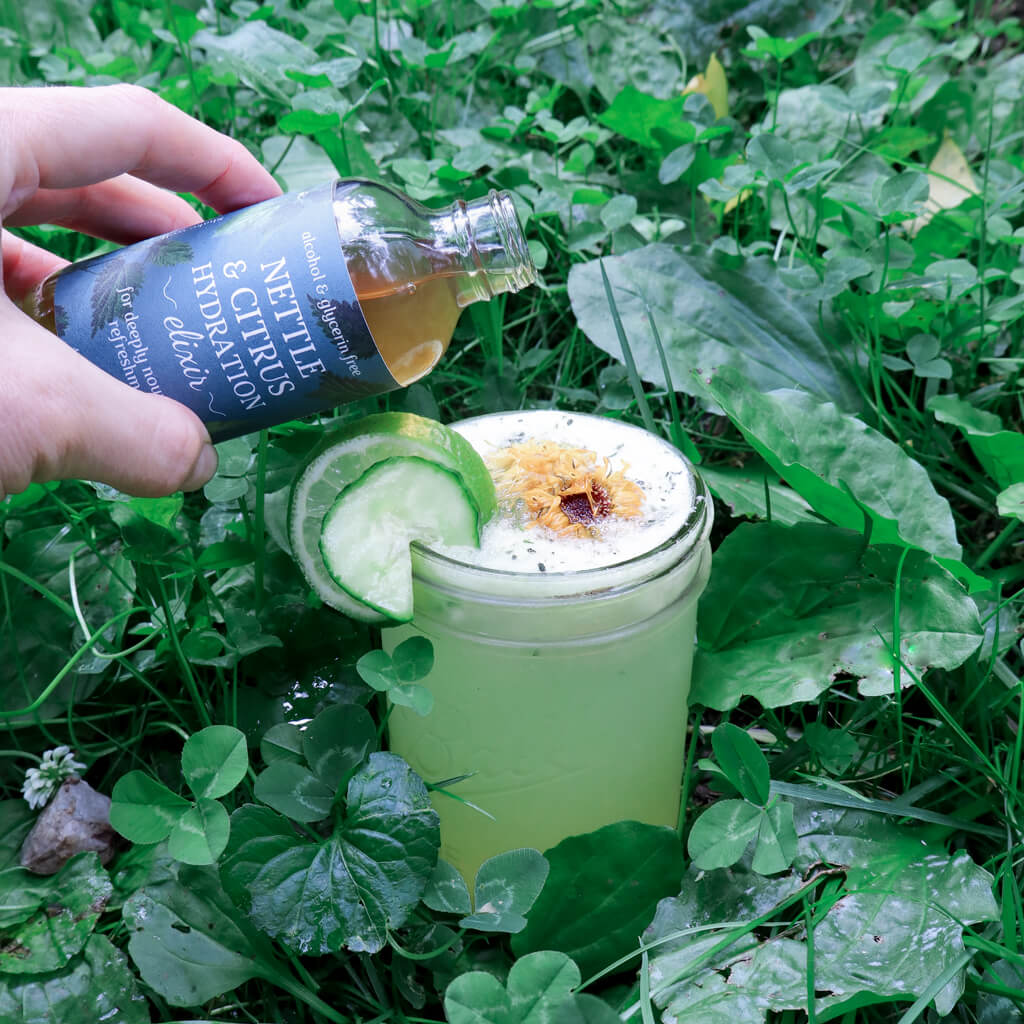 Hand pouring a bottle of Nettle & Electrolyte Elixir into a coconut water drink with lime and cucumber garnishes  in the grass