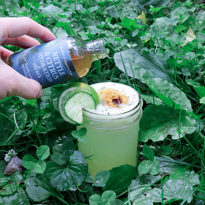 Hand pouring a bottle of Nettle & Electrolyte Elixir into a coconut water drink with lime and cucumber garnishes  in the grass