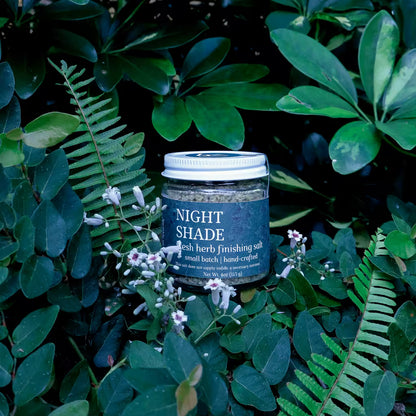 glass jar of herb infused salt with a label that reads 'Night Shade Fresh Herb Finishing Salt' in a natural setting with lush greenery and white flowers as decoration