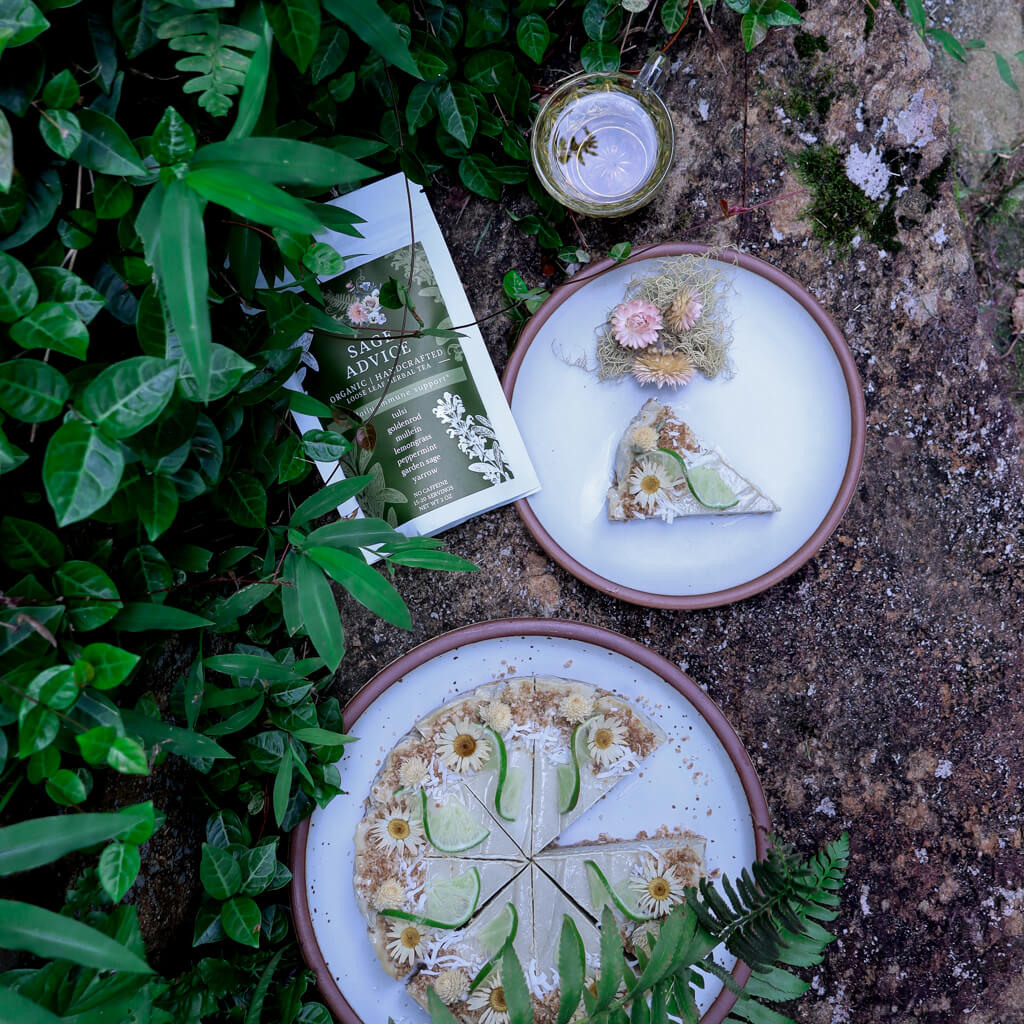 Two ceramic plates with slices of vegan lime cheesecake with a bag of Sage Advice Herbal Tea, a tea cup, and decorative elements on a natural setting with greenery.