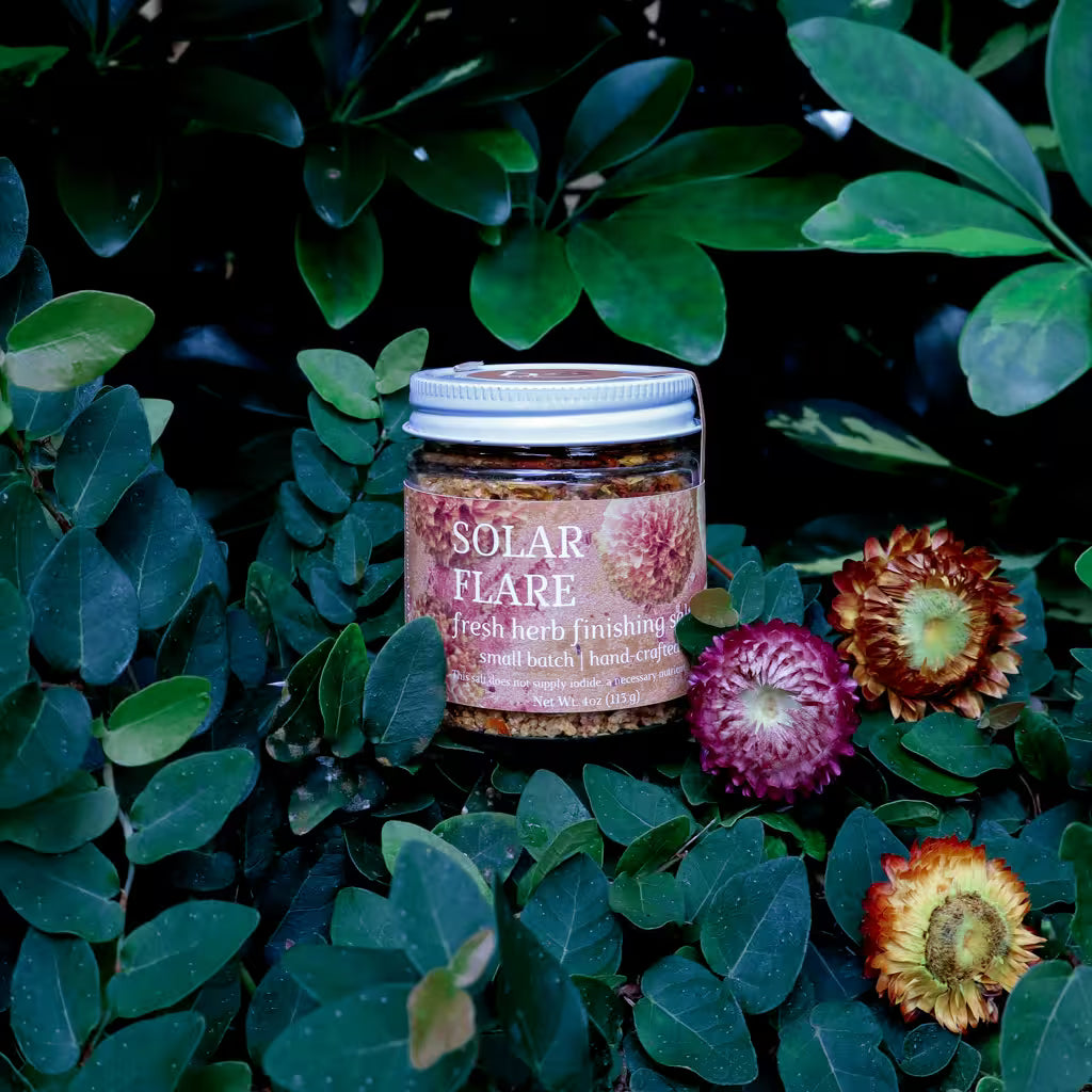 glass jar of herb infused salt with a label that reads 'Solar Flare Fresh Herb Finishing Salt' in a natural setting with lush greenery and dried flowers as decoration
