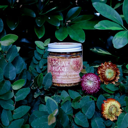 glass jar of herb infused salt with a label that reads 'Solar Flare Fresh Herb Finishing Salt' in a natural setting with lush greenery and dried flowers as decoration