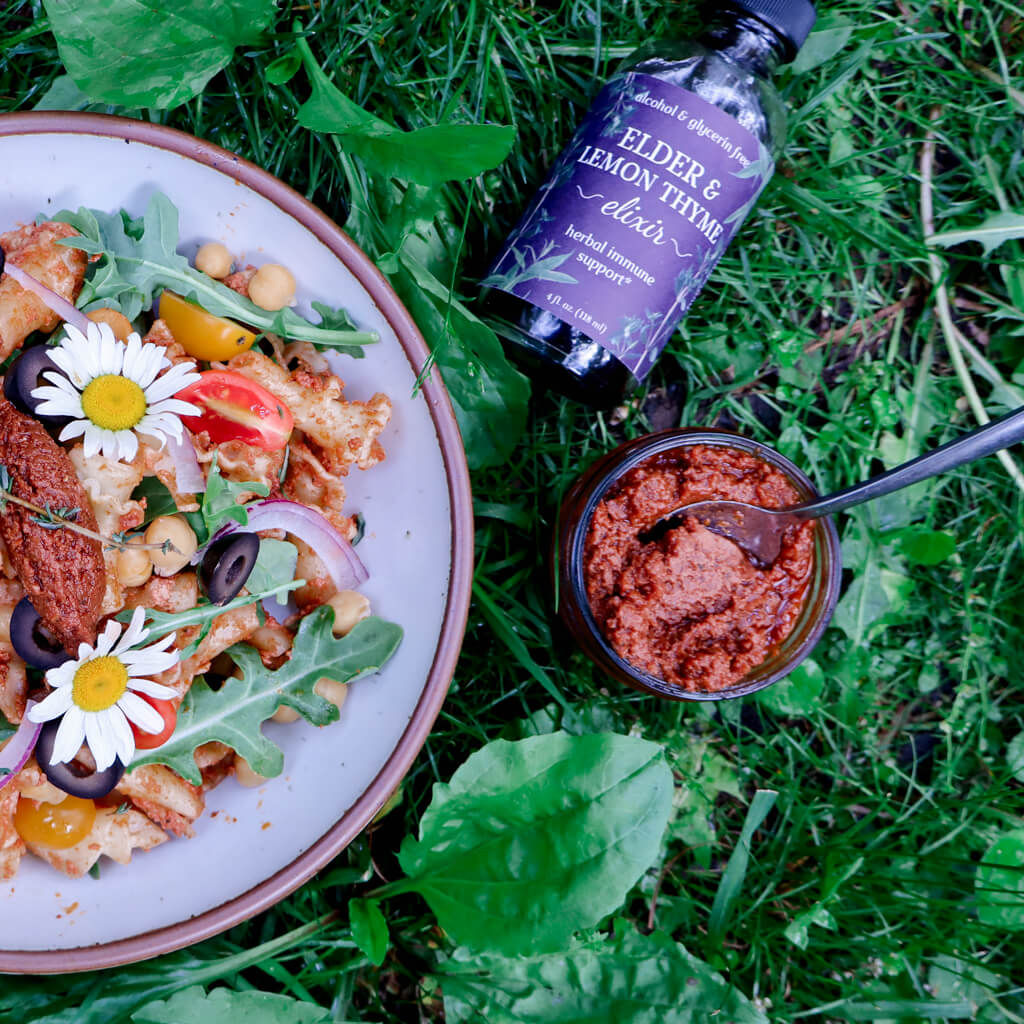 a bowl of sun-dried tomato pesto pasta salad sitting in the grass next to a ramekin of pesto and a bottle of Elder & Lemon Thyme Elixir