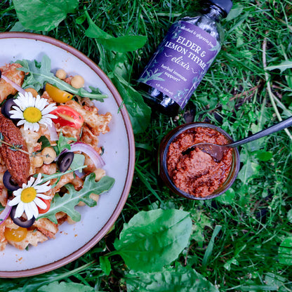 a bowl of sun-dried tomato pesto pasta salad sitting in the grass next to a ramekin of pesto and a bottle of Elder & Lemon Thyme Elixir