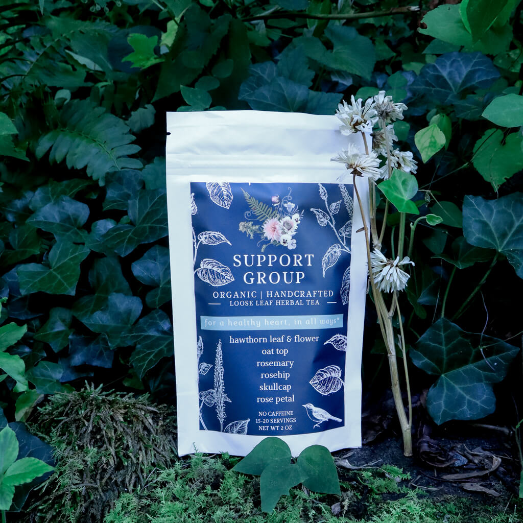 A pouch of Support Group Organic Handcrafted Loose Leaf Herbal Tea, photographed outdoors against a background of green ivy and moss with small wildflowers.