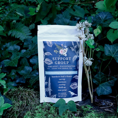 A pouch of Support Group Organic Handcrafted Loose Leaf Herbal Tea, photographed outdoors against a background of green ivy and moss with small wildflowers.