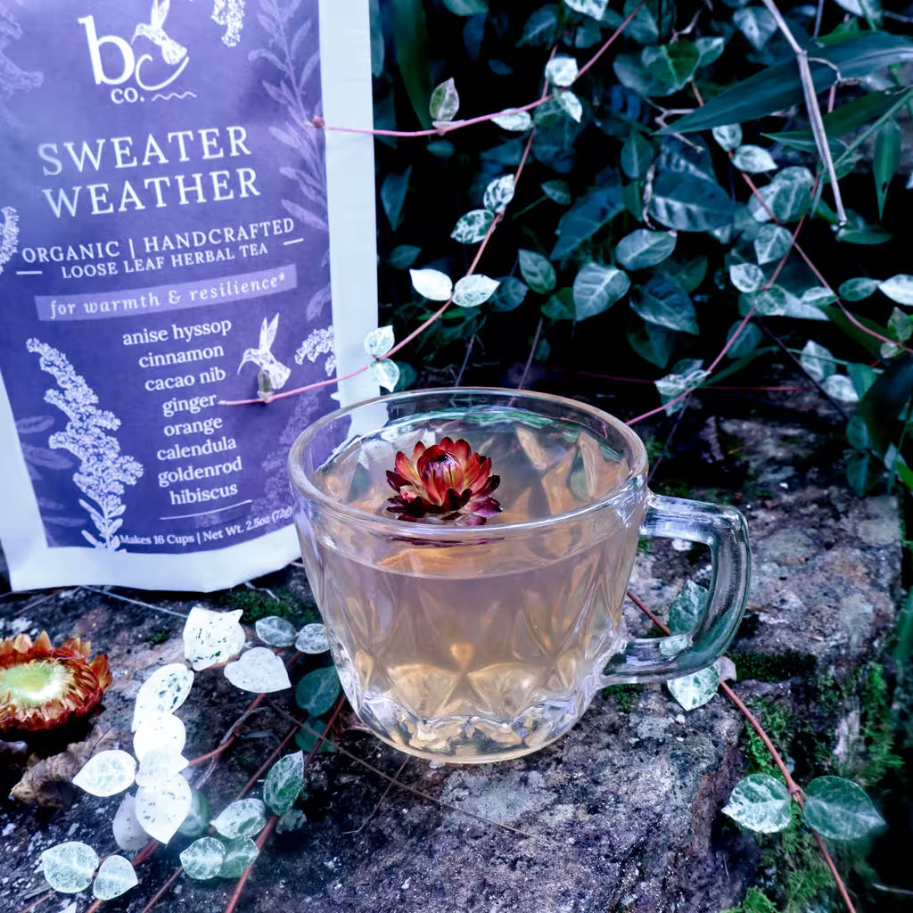A clear glass mug filled with brewed herbal tea sits on a stone surface outdoors, with a branded tea pouch labeled 'Sweater Weather' visible behind it.
