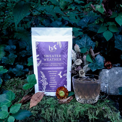 a bag of loose leaf herbal tea with a purple label that reads "Sweater Weather for warmth and resilience" in a natural setting with dried flowers as decoration and a mug of tea