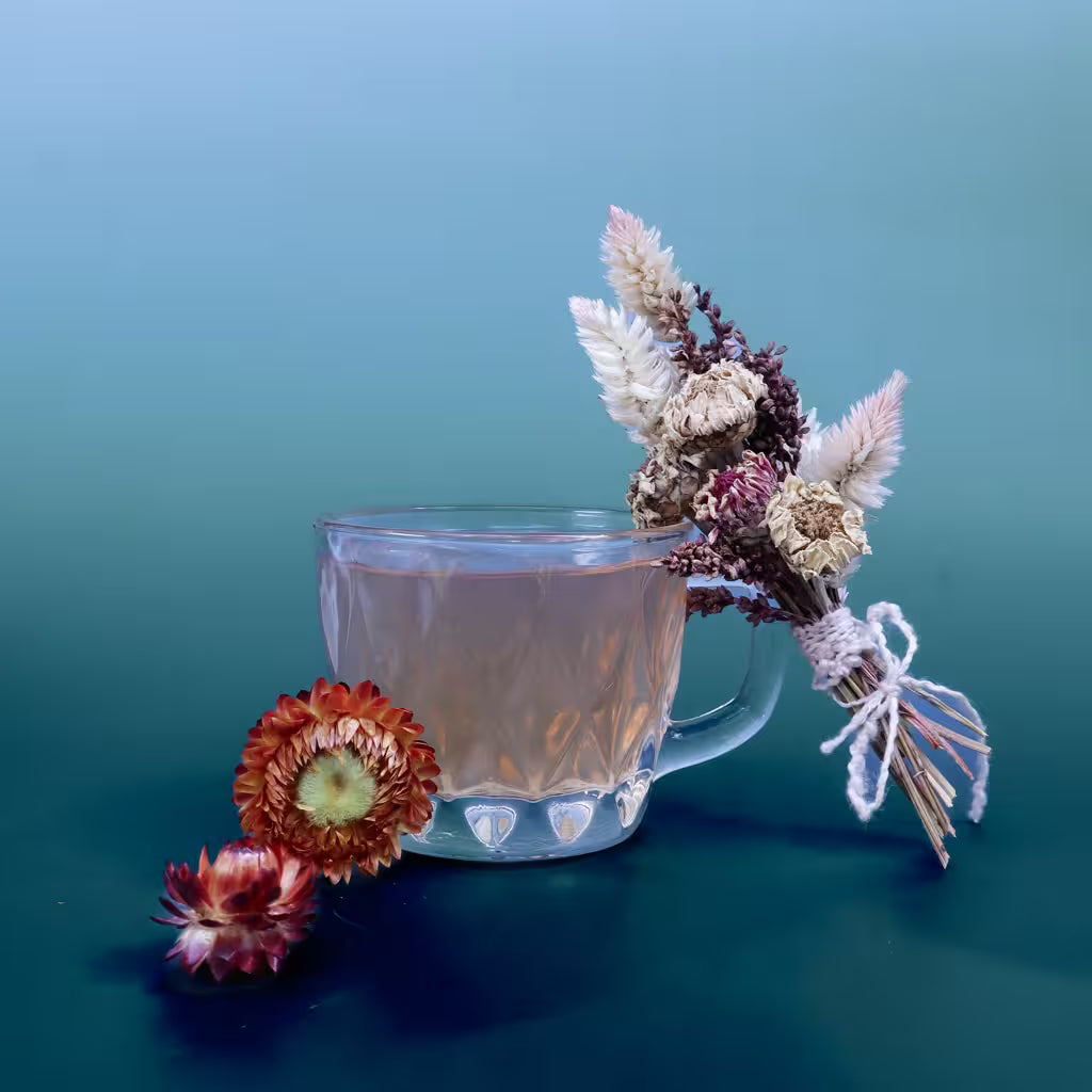 A clear glass mug filled with a pale pink herbal tea, styled with dried flowers on a blue gradient background.