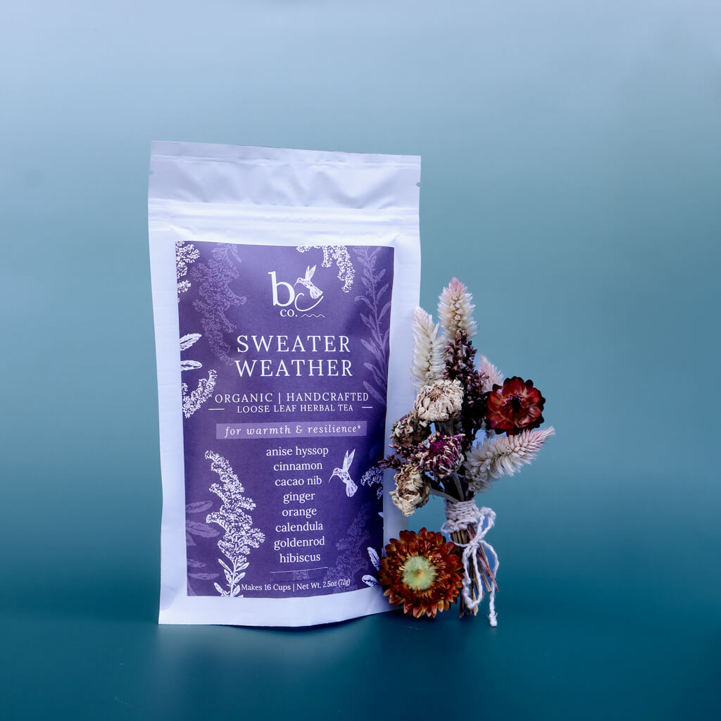 bag of "Sweater Weather Loose Leaf Organic Herbal Tea" next to a small bouquet of dried flowers against a gradient blue background