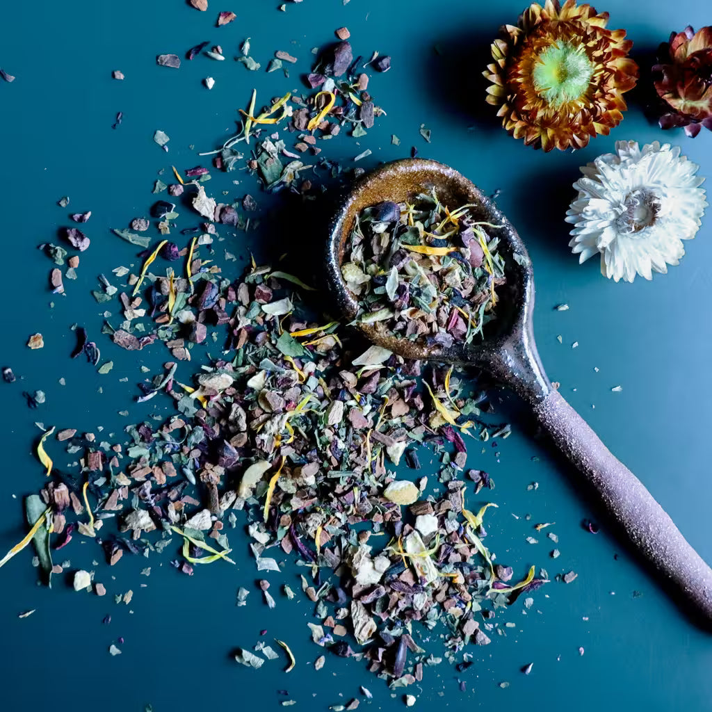 A top-down styled moody flat-lay of a warming loose-leaf herbal tea blend for fall. The tea is scattered across a dark teal surface with a rustic ceramic spoon filled with the dried tea blend and dried flower decor.
