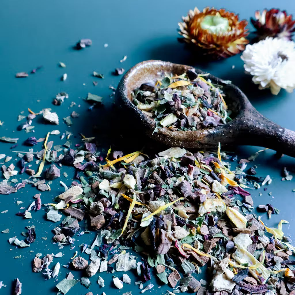A rustic ceramic spoon filled with loose-leaf herbal tea sits on a dark teal surface, with scattered dried tea pieces spilled around it. The tea blend contains anise hyssop, cinnamon, cacao nibs, orange peel, ginger, and calendula.