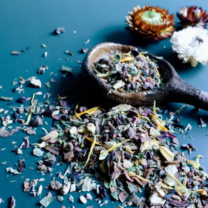 A rustic ceramic spoon filled with loose-leaf herbal tea sits on a dark teal surface, with scattered dried tea pieces spilled around it. The tea blend contains anise hyssop, cinnamon, cacao nibs, orange peel, ginger, and calendula.